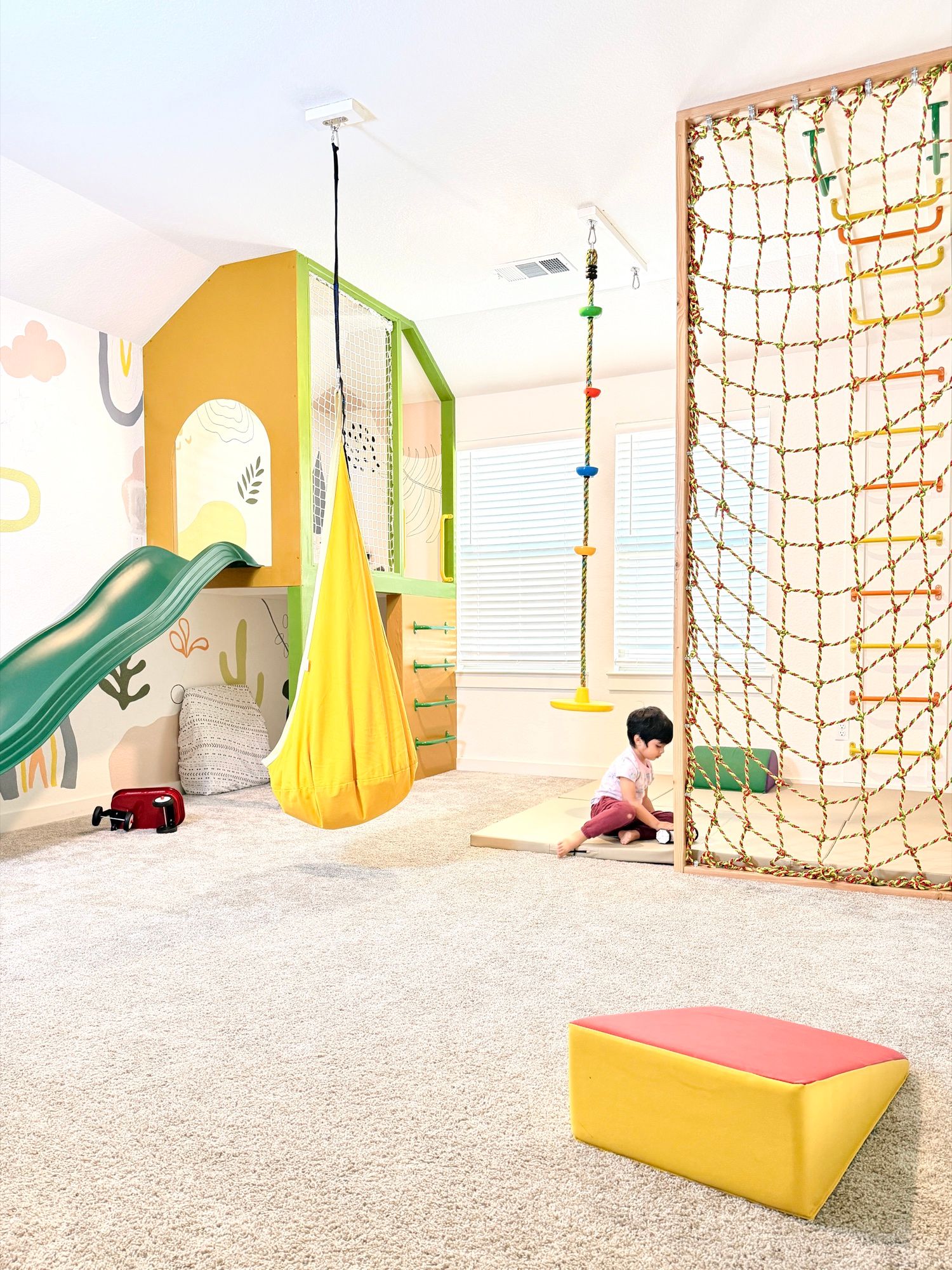 The Sunshine House: a sunshine-yellow playhouse with a green slide, a pod swing and a rope net