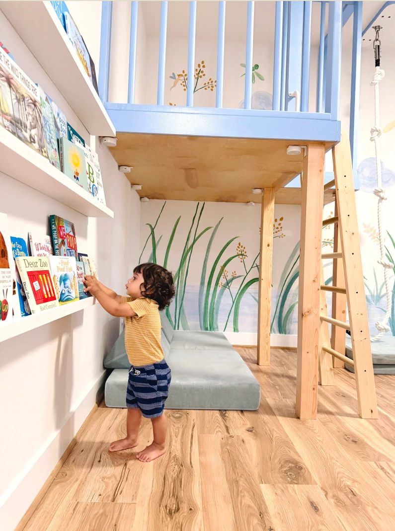 A toddler choosing a book from wall ledges beside a cushioned nook under a blue loft