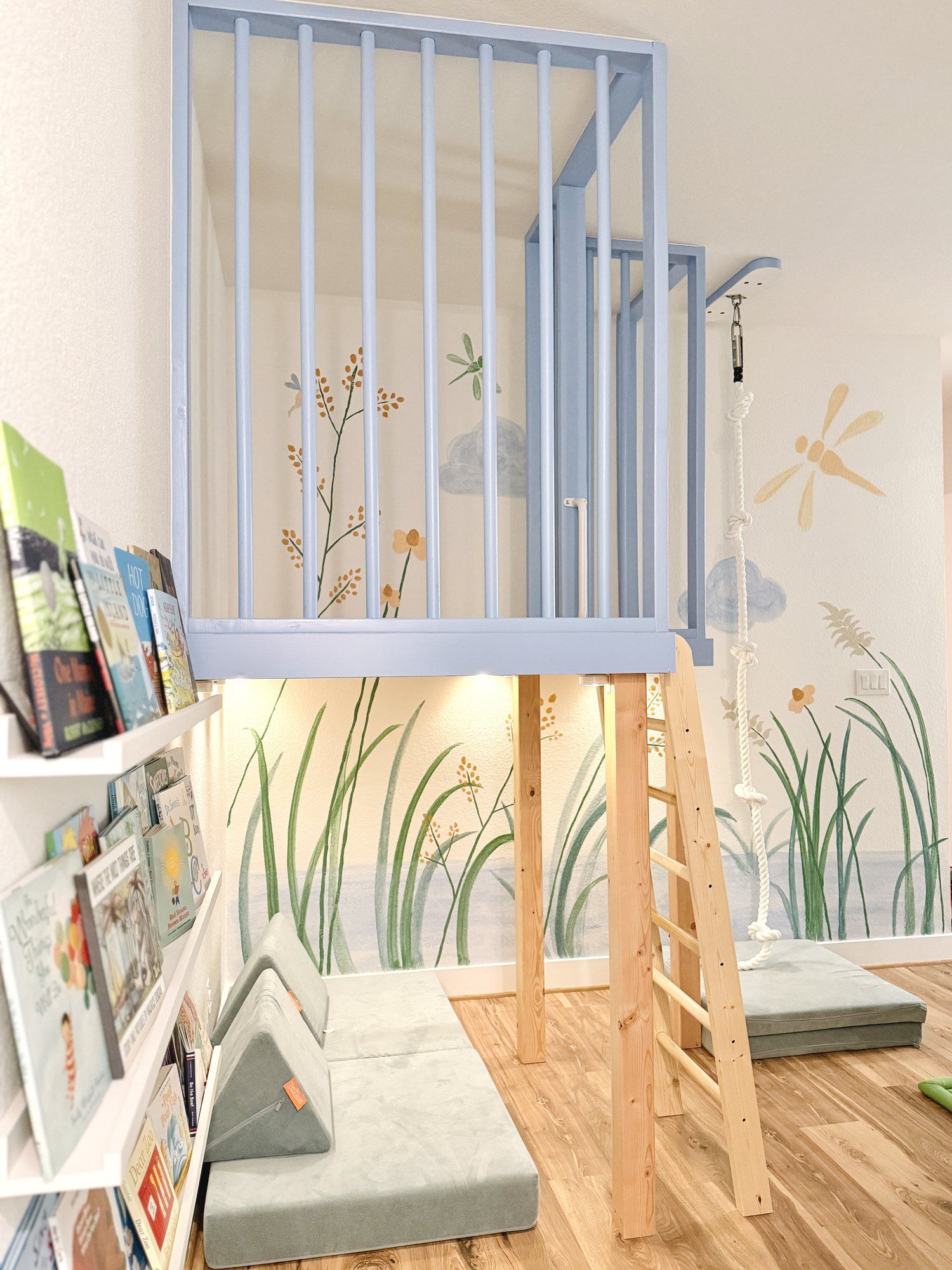 A soft blue play loft with a wooden ladder and a reading corner beneath, beside a meadow mural