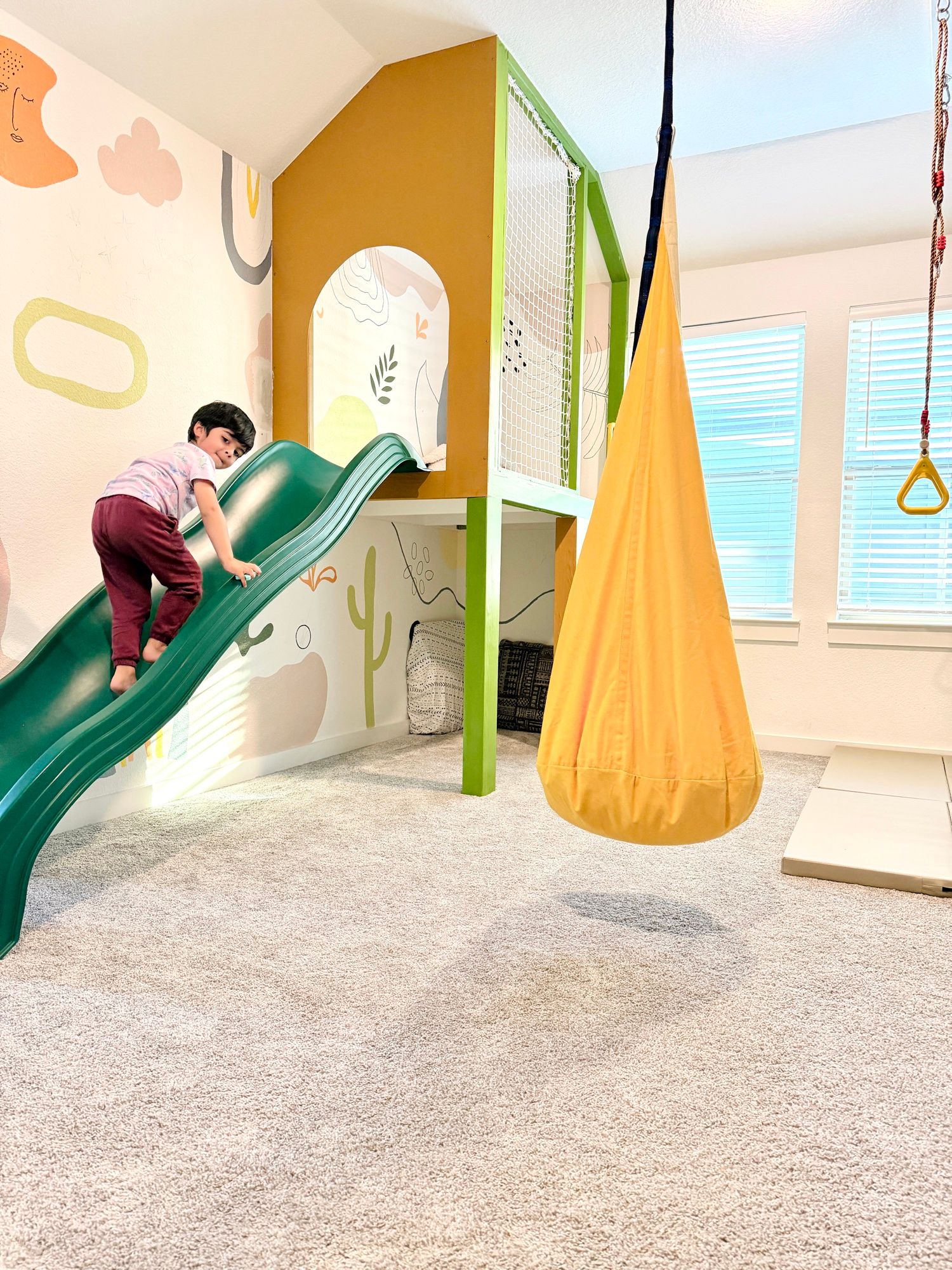 A child climbing up the green slide beside the yellow pod swing