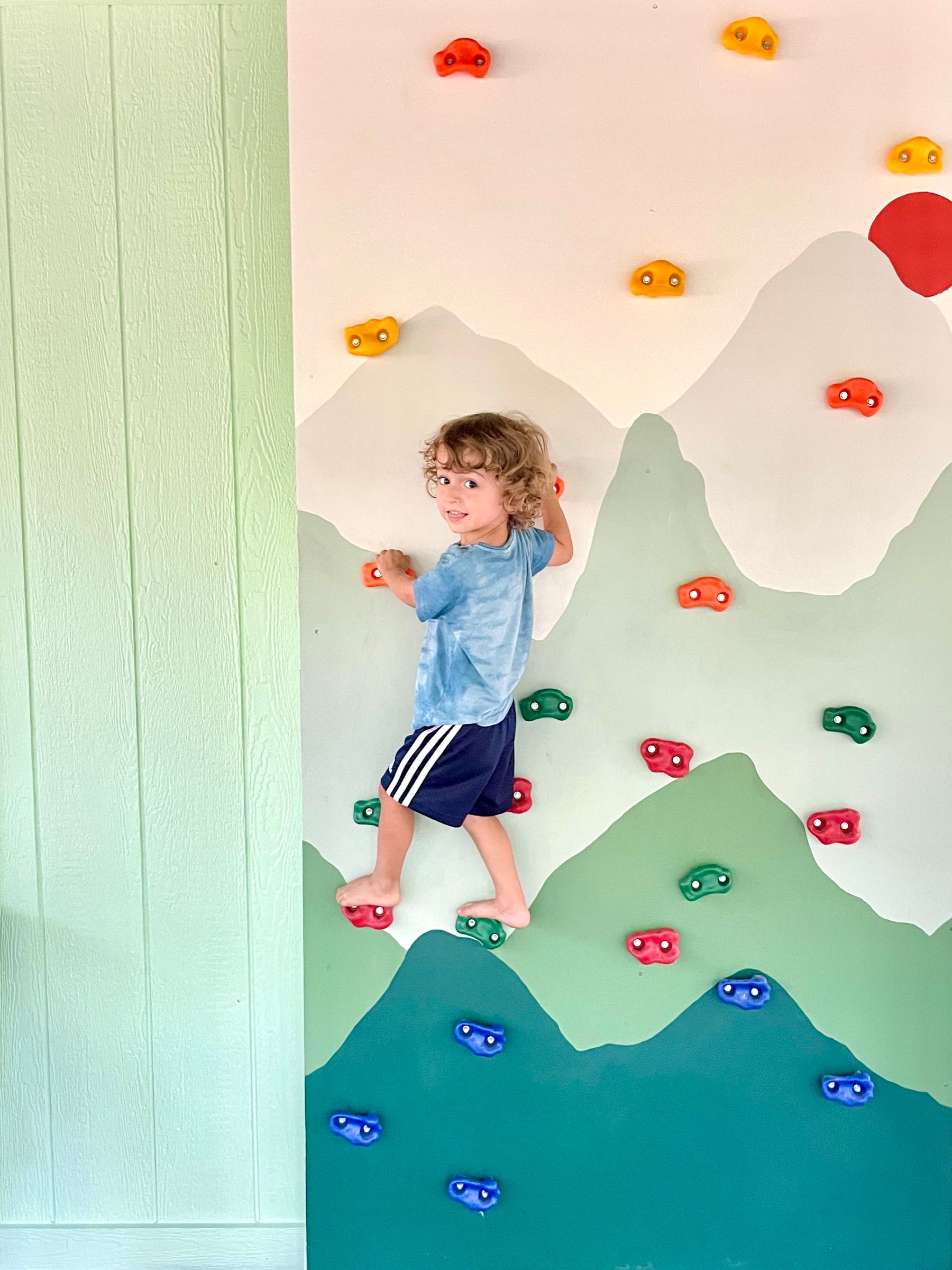 A child looking back over their shoulder while climbing a wall painted with green mountains