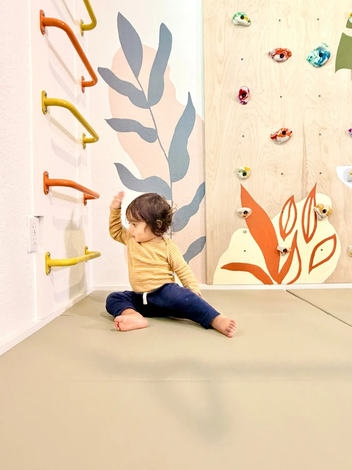 A toddler sitting on the soft mats of a Movement Nook, reaching up towards the colourful monkey bars beside a climbing wall painted with leaves