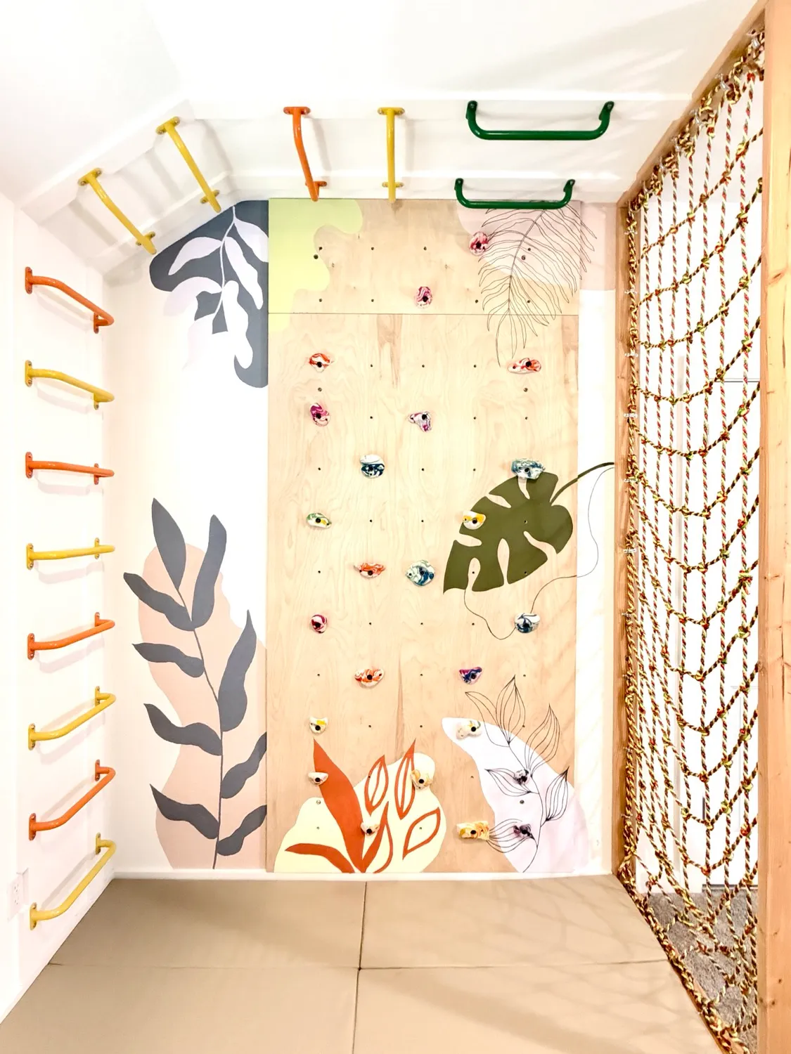 A Movement Nook with a plywood climbing wall, colourful monkey bars, a rope climbing net and soft mats