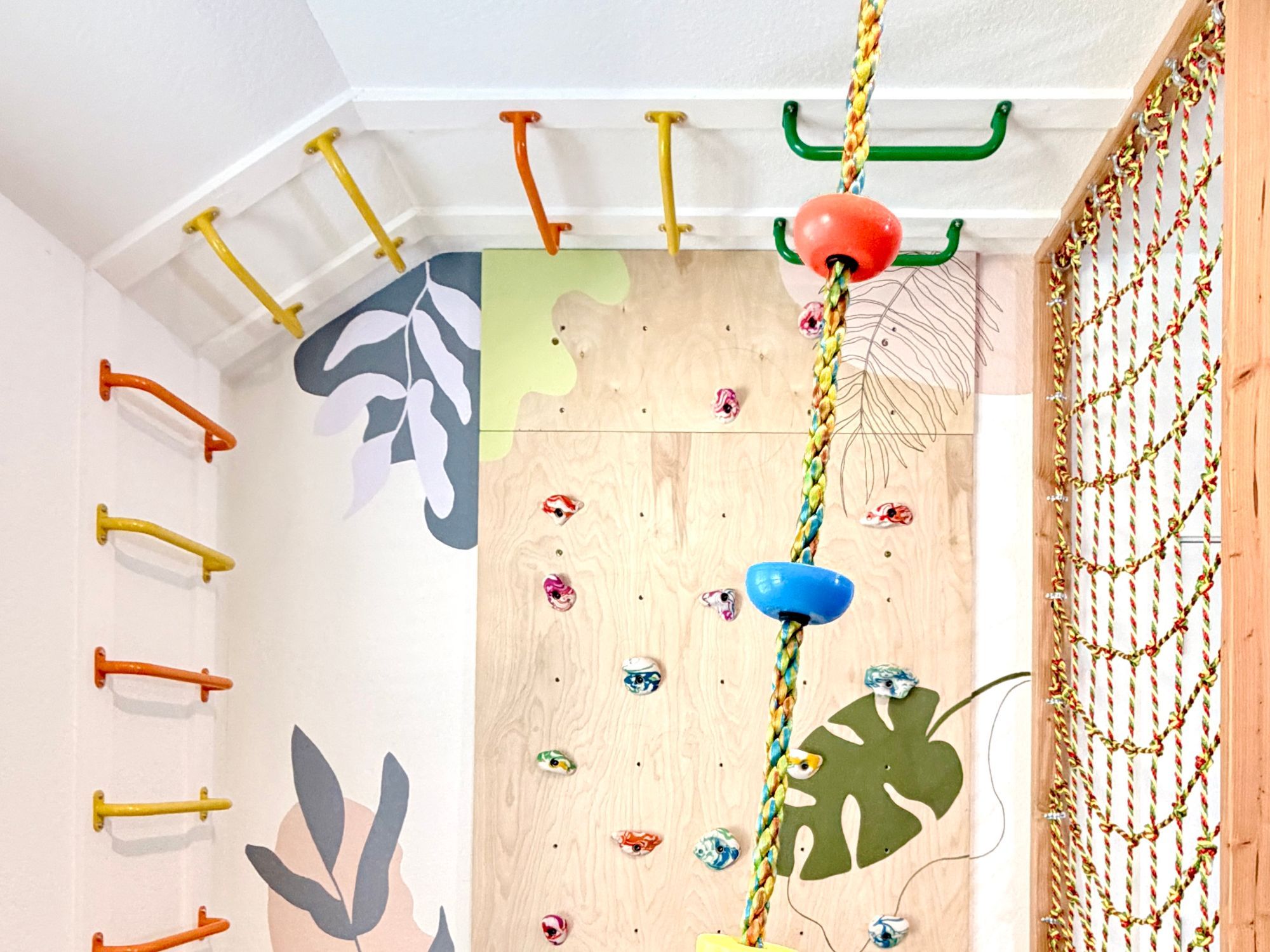 A Movement Nook: colourful monkey bars along the wall and ceiling, a plywood climbing wall painted with leaves, a disc-swing rope and a rope net