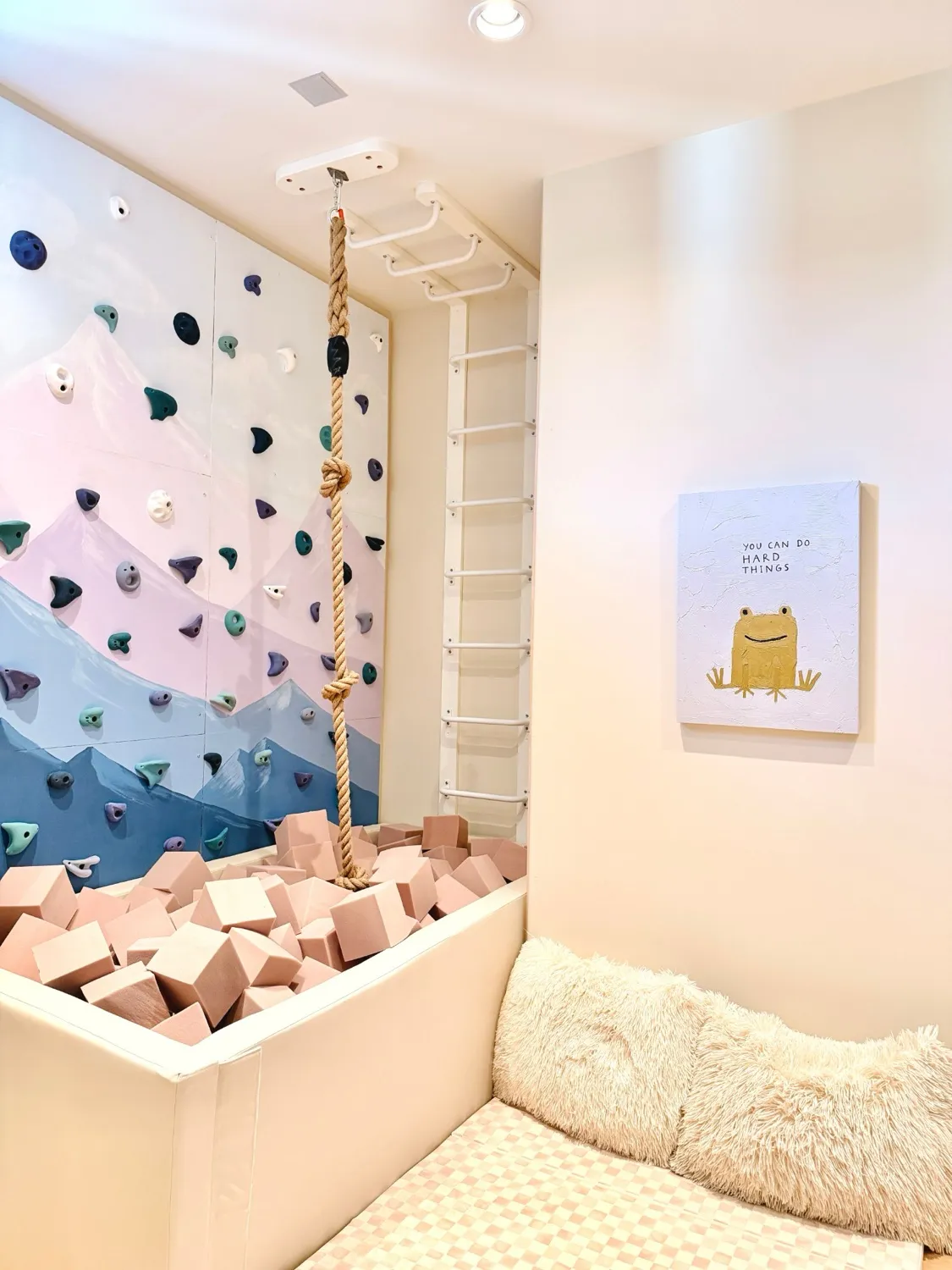 A painted mountain climbing wall above a foam pit with a knotted rope and monkey bars, beside a cosy corner with fluffy cushions and a frog picture saying You can do hard things