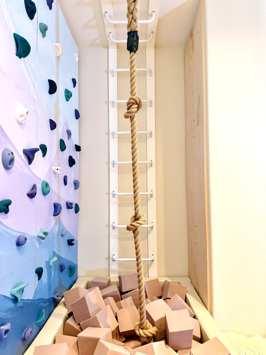 A knotted climbing rope hanging into a foam pit beside a ladder of monkey bars and the climbing wall