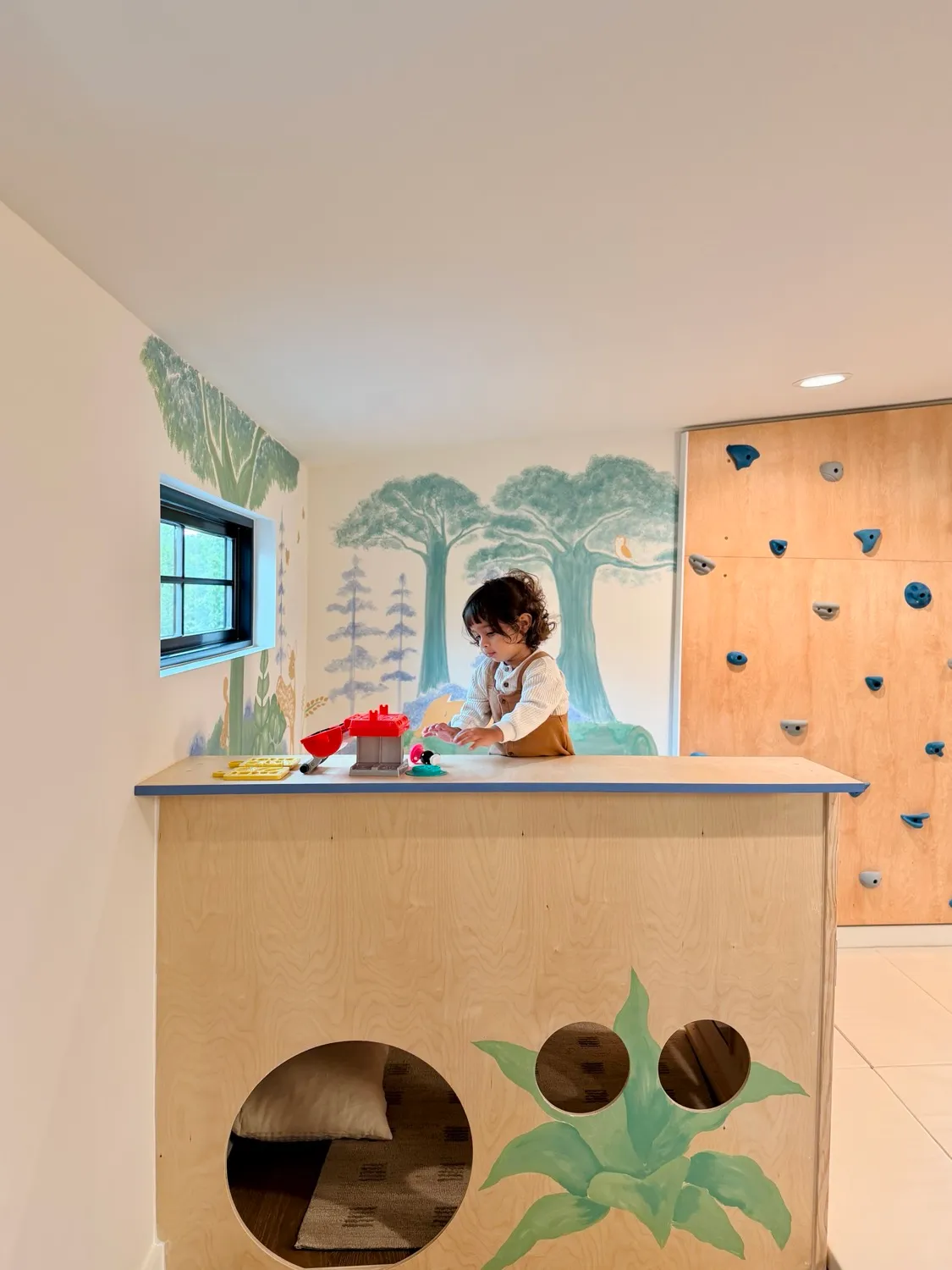 A child playing at the counter of the low wooden platform, with round windows into the den below and a climbing wall behind