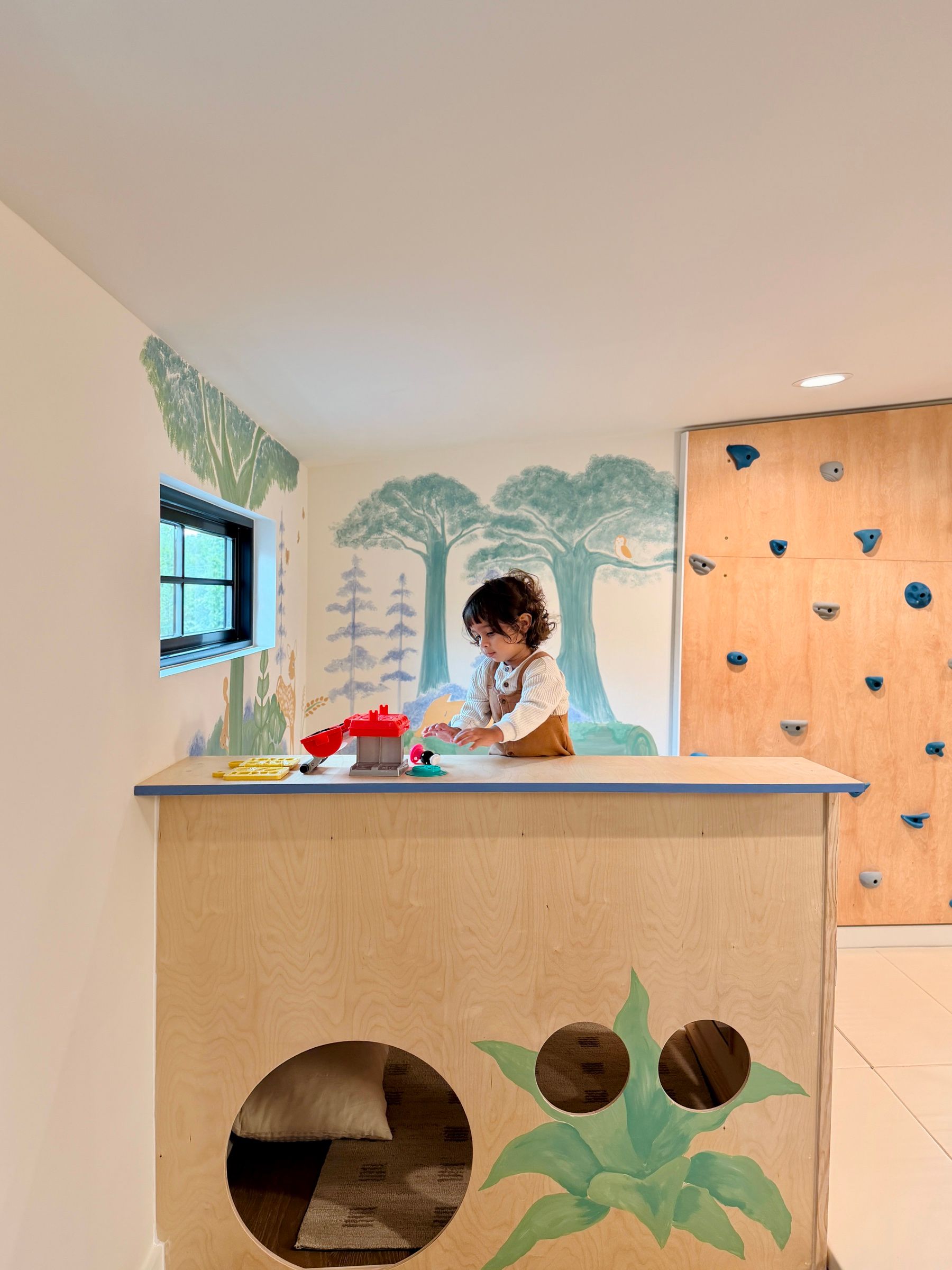 A child playing at the counter of a low wooden platform, with a painted woodland mural and a climbing wall behind