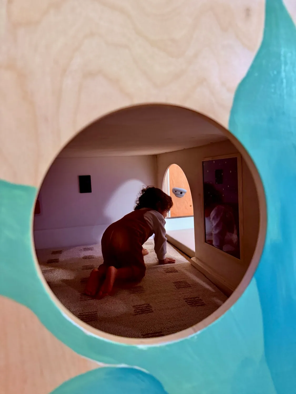 A child crawling into the carpeted den beneath the platform, seen through a round window