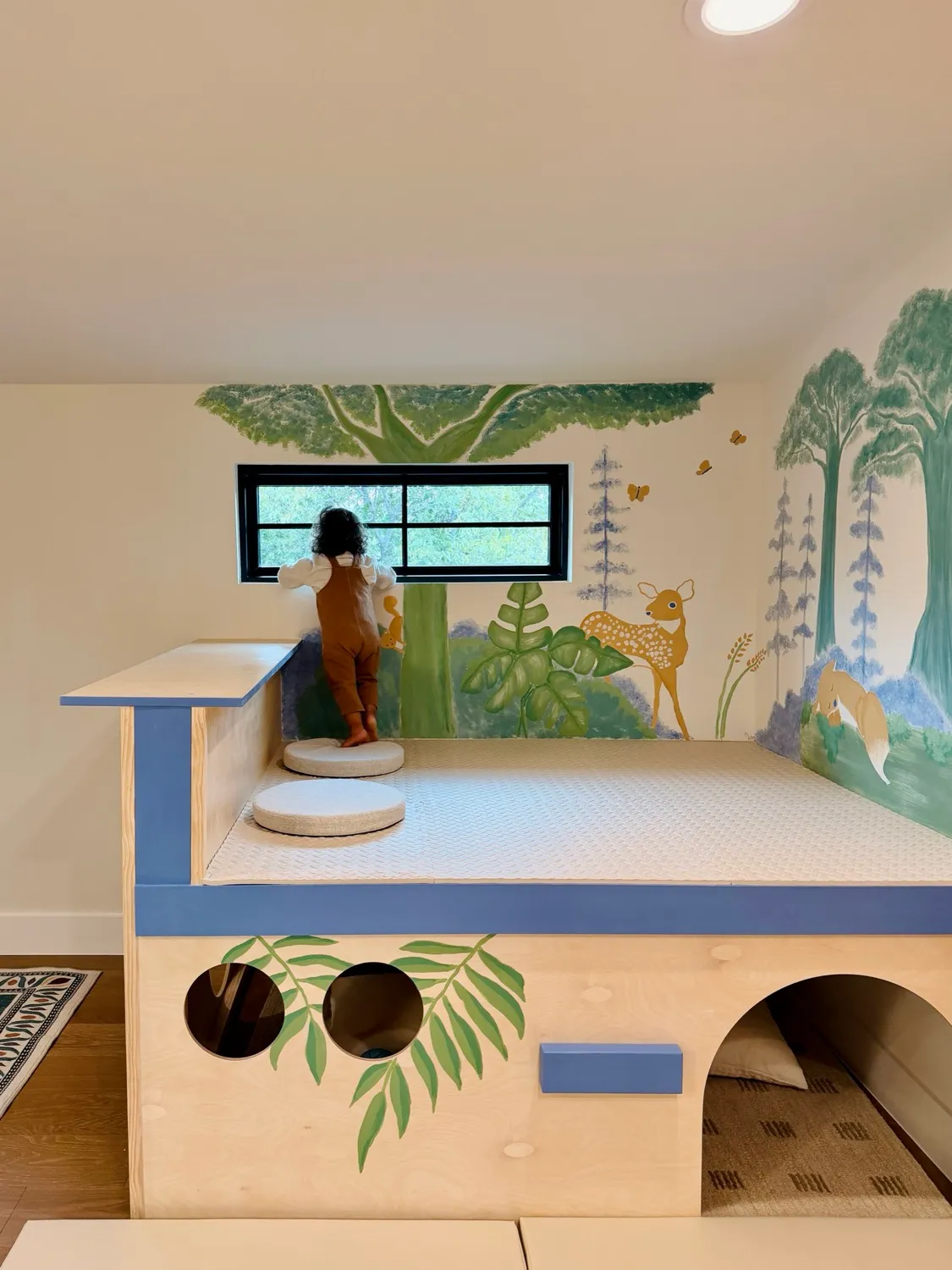 A child at the window on a low wooden platform with a cushioned top and a den with round windows beneath, under a painted woodland mural